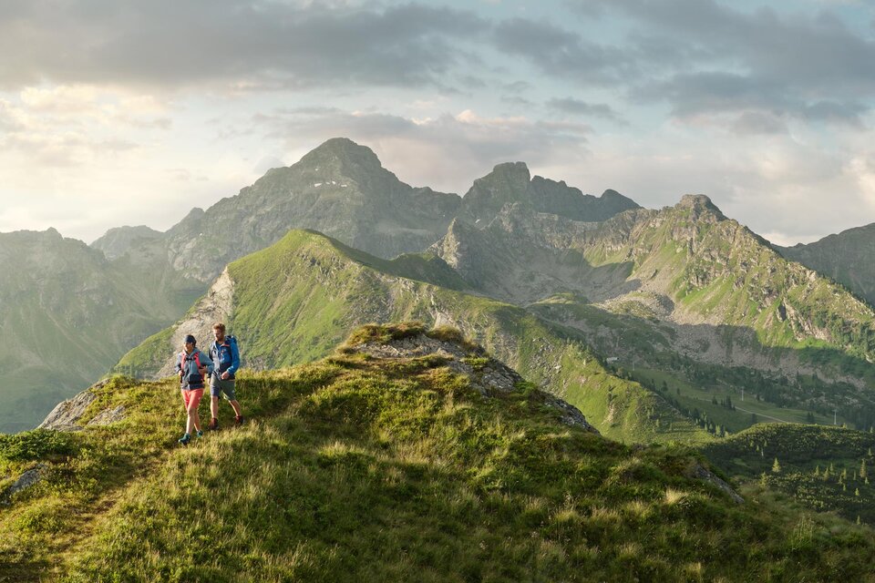 Ein atemberaubender Blick auf die Berge mit üppigem Gras im Vordergrund. Zwei Personen stehen auf einem Gipfel und genießen die Landschaft.