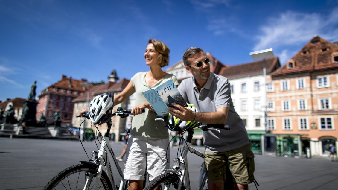 Ein Paar steht mit Fahrrädern auf einem Platz und schaut auf eine Karte. Die Sonne scheint und im Hintergrund sind schöne Gebäude zu sehen. | © Graz Tourismus-Tom Lamm