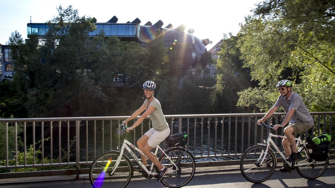 Zwei Radfahrer fahren auf einem Weg entlang eines Gewässers. Die Sonne scheint und es ist eine grüne Umgebung zu sehen. | © Graz Tourismus-Tom Lamm