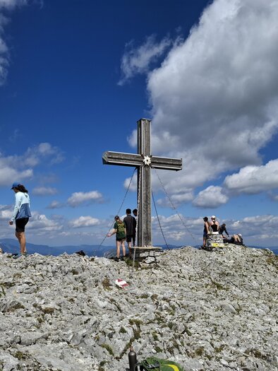 Eine Gruppe von Wanderern steht auf einem felsigen Gipfel neben einem großen Kreuz. Der Himmel ist blau mit weißen Wolken und es ist ein sonniger Tag. | © Michael Geißler