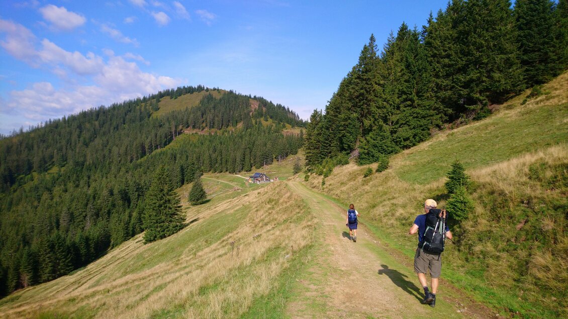 Zwei Wanderer gehen einen Pfad entlang, umgeben von grünen Wäldern und Bergen. Der Himmel ist klar mit einigen Wolken. | © Michael Geißler