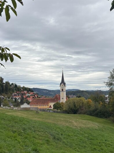 Blick über eine grüne Wiese auf die Wallfahrtskirche Maria Lankowitz mit ihrem Kirchturm, umgeben von Häusern und Hügeln. | © Kohlbacher