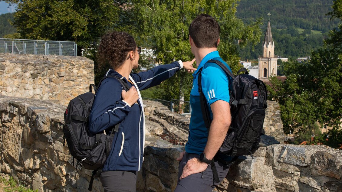 Zwei Wanderer stehen am Rand einer Steinmauer und schauen auf eine Kirche in der Ferne. Die Umgebung ist grün mit Bäumen und sanften Hügeln. | © TVB Erzberg-Leoben