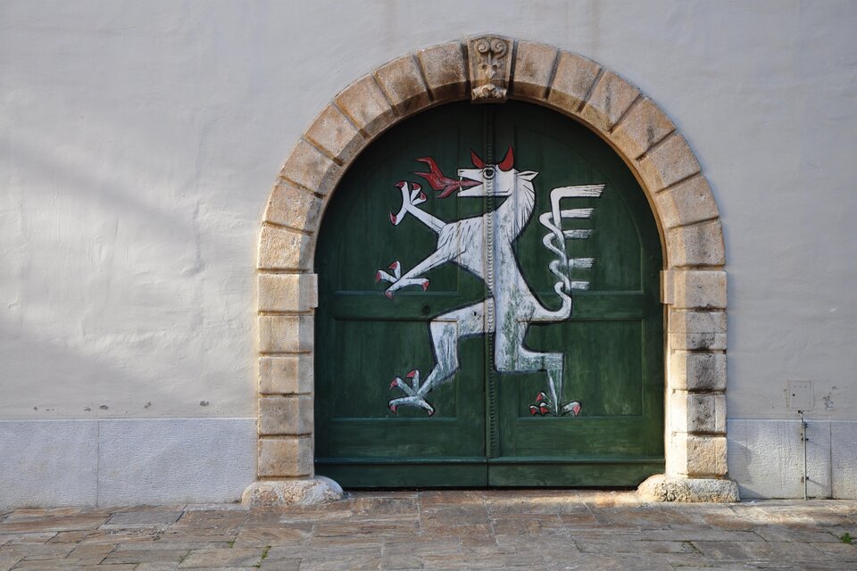 Ein grünes Tor mit einem gemalten, weißen, mythischen Wesen. Das Tor hat einen runden Steinrahmen und befindet sich an einer hellen Wand. | © (c) Daniel Strohrigl