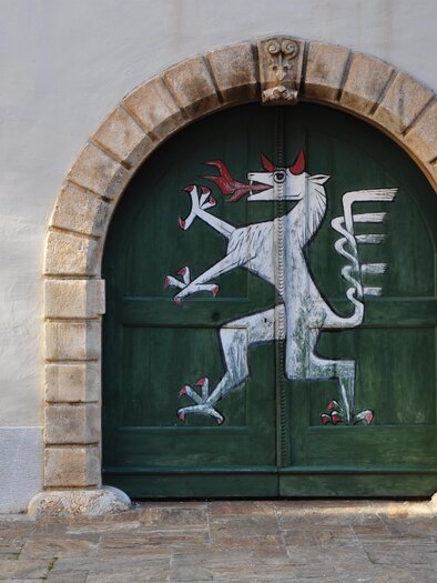 A green gate with a painted white mythical creature. The gate has a round stone frame and is situated on a light wall. | © (c) Daniel Strohrigl