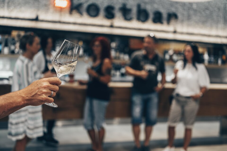 Eine Person hält ein Glas mit Getränk in der Hand, während im Hintergrund mehrere Menschen an einer Bar stehen. Die Atmosphäre ist gesellig und einladend. | © Ian Ehm