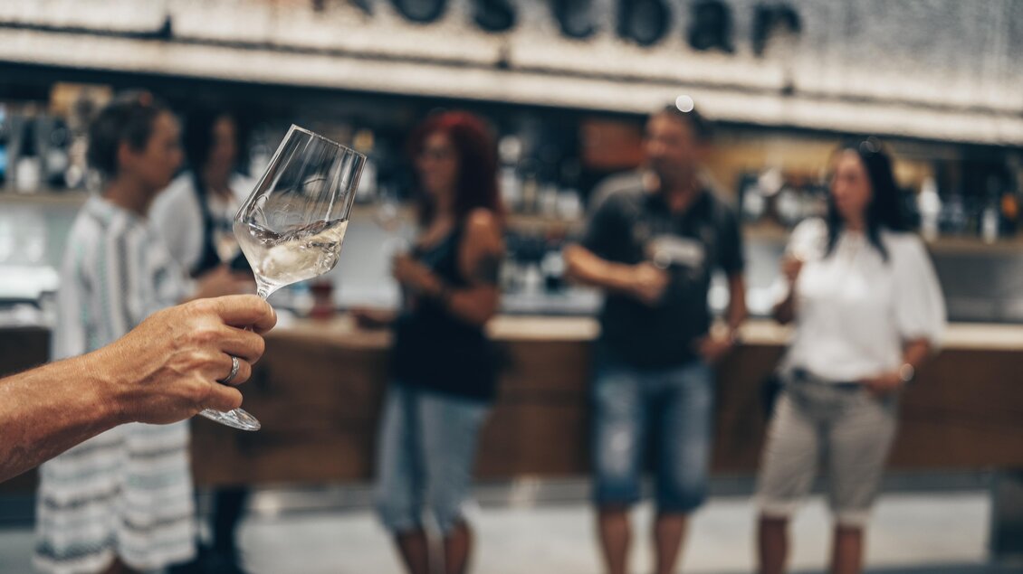 Eine Person hält ein Glas mit Getränk in der Hand, während im Hintergrund mehrere Menschen an einer Bar stehen. Die Atmosphäre ist gesellig und einladend. | © Ian Ehm