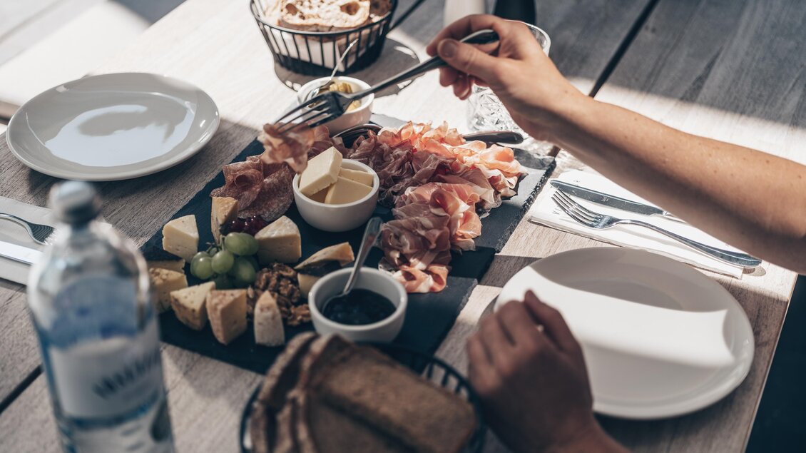 Ein leckeres Buffet mit verschiedenen Käsesorten, Schinken und Trauben auf einem Tisch. Im Hintergrund sind Teller und eine Flasche Wasser zu sehen. | © Ian Ehm