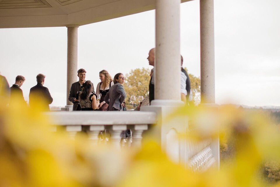 Eine Gruppe von Menschen steht unter einem Pavillon und unterhält sich. Im Vordergrund sind gelbe Blätter sichtbar, die die herbstliche Stimmung unterstreichen. | ©  Karl Schrotter