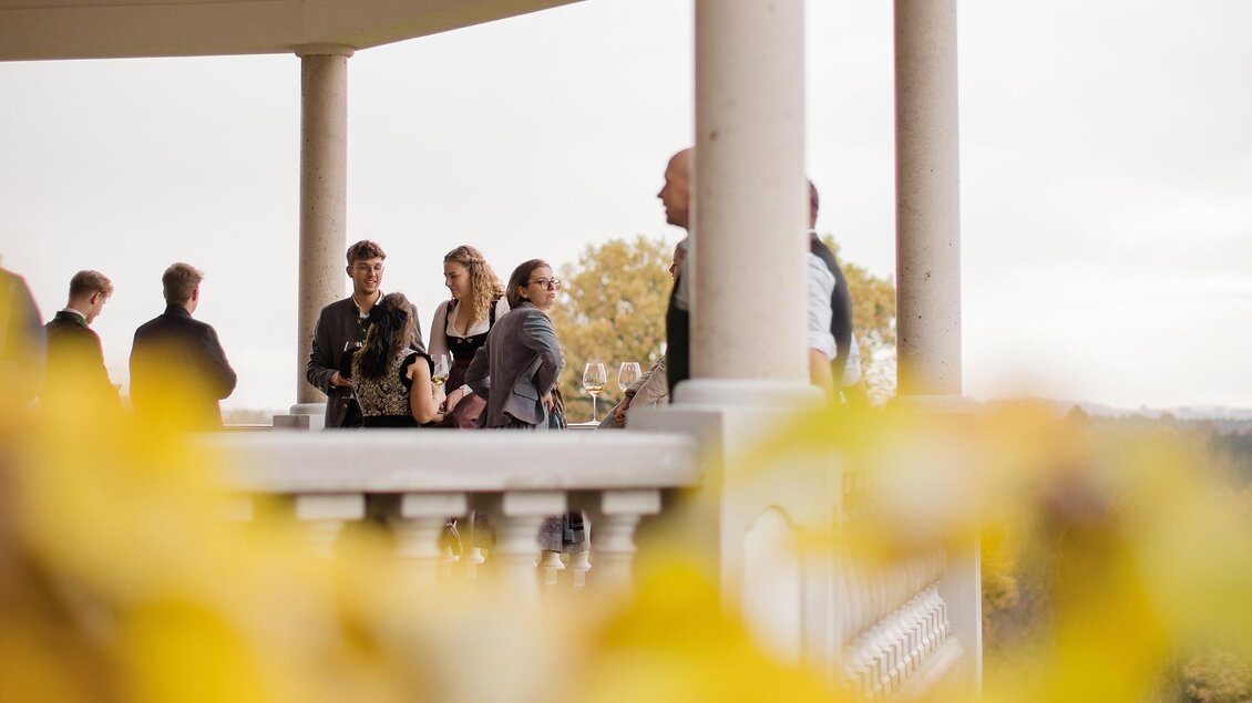 Eine Gruppe von Menschen steht unter einem Pavillon und unterhält sich. Im Vordergrund sind gelbe Blätter sichtbar, die die herbstliche Stimmung unterstreichen. | ©  Karl Schrotter