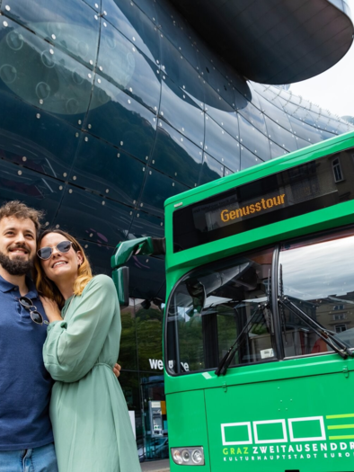 A couple is smiling in front of a green bus ticket, while a modern building can be seen in the background. The surroundings feel vibrant and inviting.