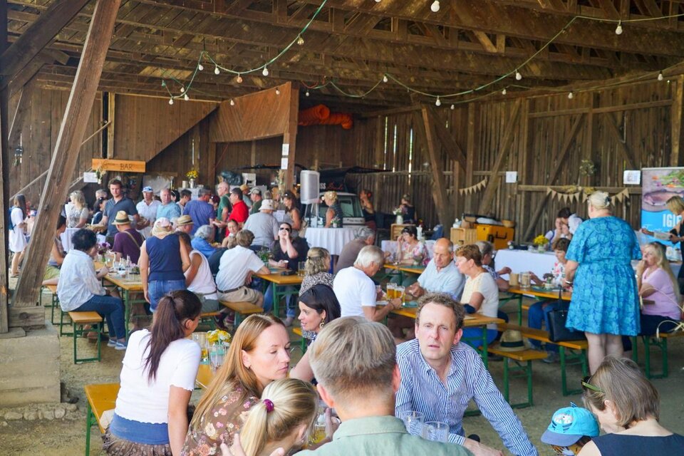 Eine lebhafte Veranstaltung in einer Scheune mit vielen Menschen, die an Tischen sitzen und sich unterhalten. Die Atmosphäre ist gesellig und einladend. | © GenussReich