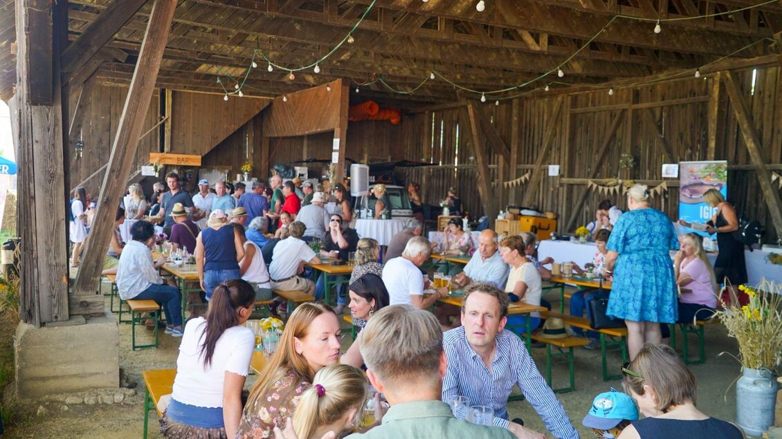 Eine lebhafte Veranstaltung in einer Scheune mit vielen Menschen, die an Tischen sitzen und sich unterhalten. Die Atmosphäre ist gesellig und einladend. | © GenussReich