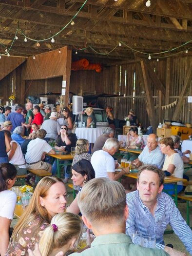 A lively event in a barn with many people sitting at tables and chatting. The atmosphere is sociable and inviting. | © GenussReich