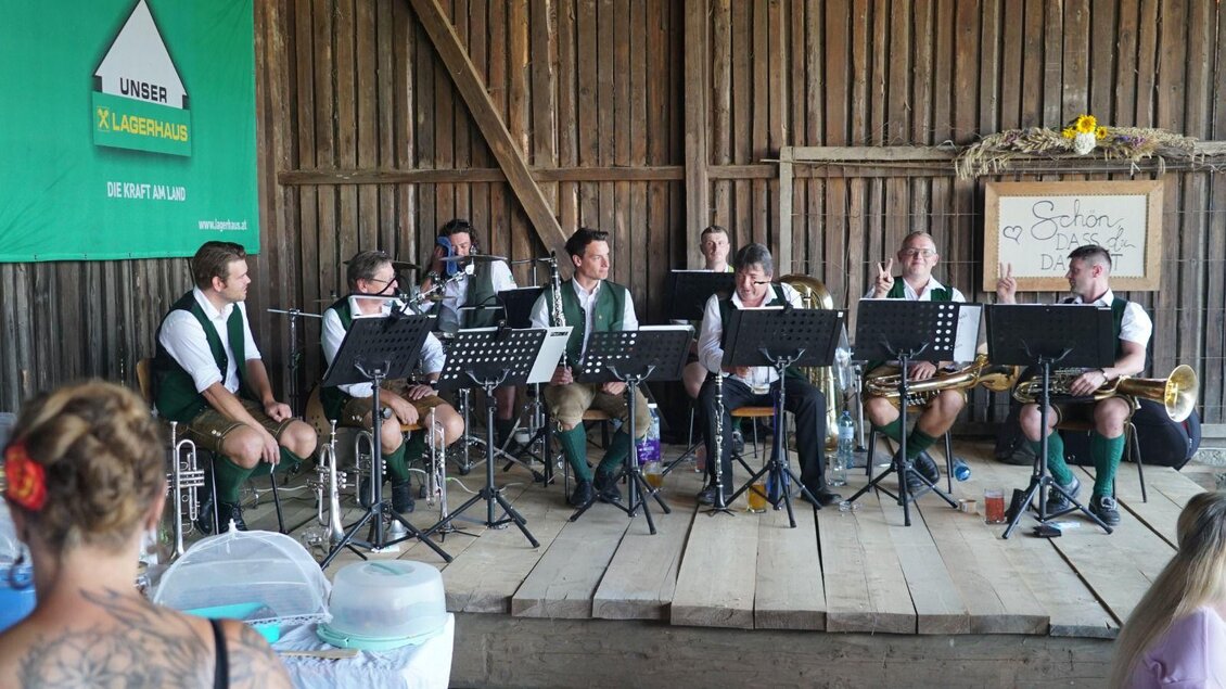 Eine Blaskapelle spielt in einer Scheune. Die Musiker tragen traditionelle Trachten und sind von Notenständern umgeben. | © GenussReich
