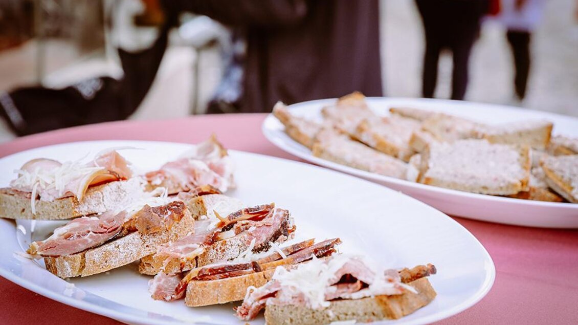 Ein Teller mit belegten Broten, garniert mit Schinken und Käse. Im Hintergrund sind weitere Brote auf einem Tablett sichtbar. | © Foto Freisinger