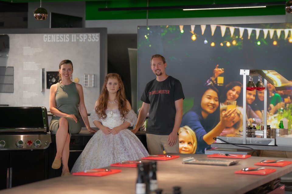 Eine Gruppe von drei Personen steht in einer modernen Küche. Im Hintergrund sind festliche Lichter und ein Familienfoto zu sehen. | © Bernd Widmoser