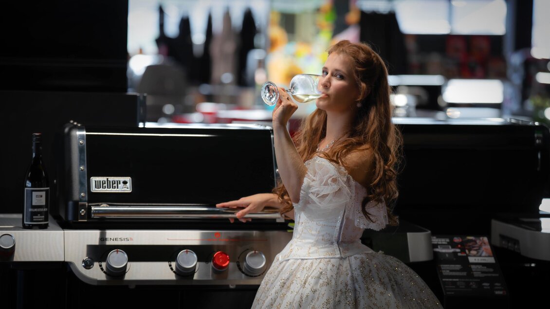 Eine Frau in einem eleganten weißen Kleid trinkt aus einem Glas neben einem Grill. Die Szene spielt in einem modernen Geschäft mit Schaufenstern im Hintergrund. | © Bernhard Widmoser