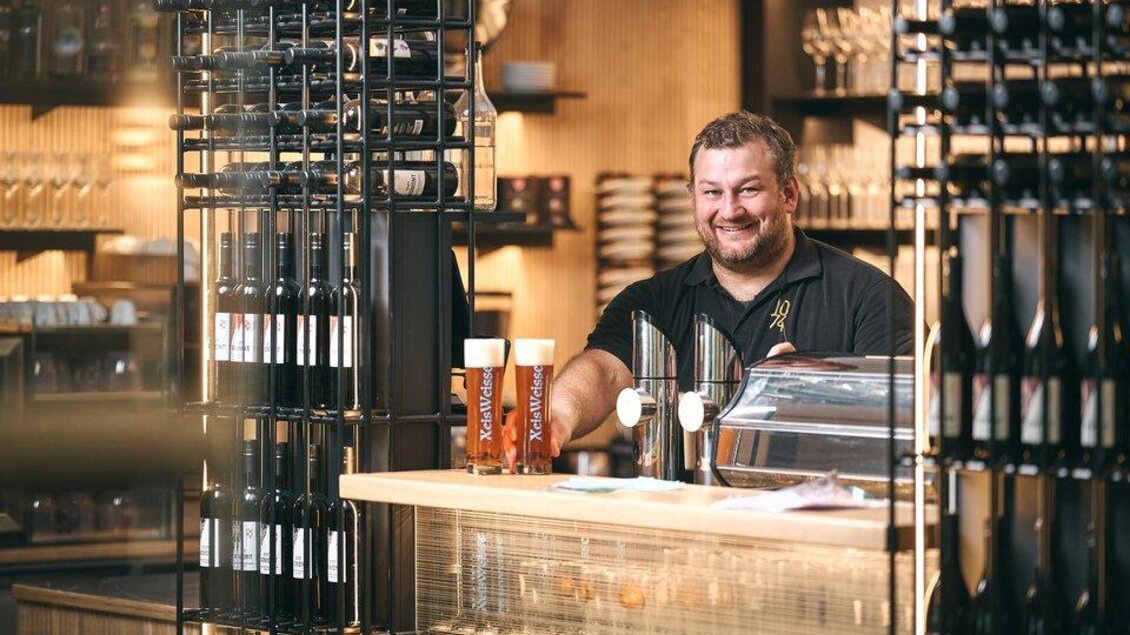 Ein freundlicher Barkeeper steht hinter der Theke in einer gemütlichen Bar. Im Hintergrund sind Weinflaschen und Gläser sichtbar. | © Gesäuse Partner