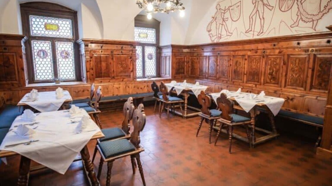 Ein gemütlicher Speisesaal mit Holzmöbeln und Tischen, die mit weißen Tischdecken gedeckt sind. Die Wände sind mit Holzvertäfelungen und historischen Zeichnungen dekoriert.