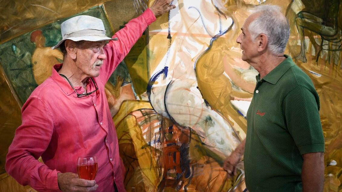 Zwei Männer unterhalten sich lebhaft vor einem Gemälde. Der Mann links trägt ein rotes Hemd und hält ein Glas in der Hand, während der andere in einem grünen Polo stark interessiert wirkt. | © Ulrike Rauch