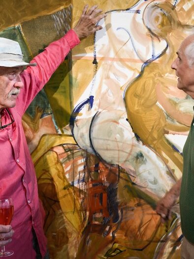 Two men are engaging in a lively conversation in front of a painting. The man on the left is wearing a red shirt and holding a glass in his hand, while the other man in a green polo appears to be very interested. | © Ulrike Rauch