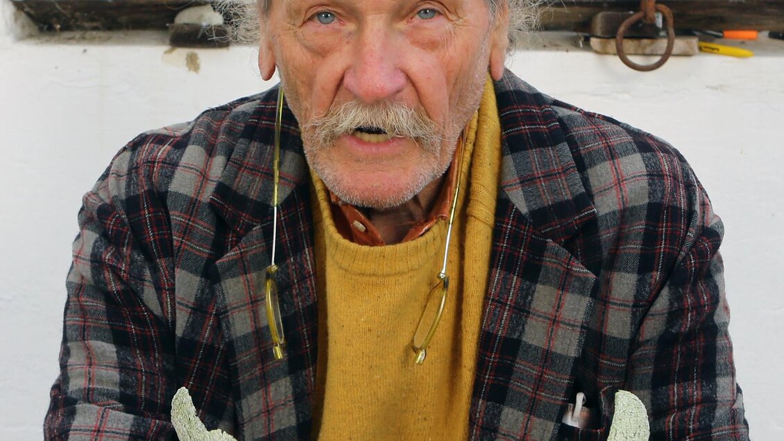 Ein älterer Mann sitzt mit einem grünen Kunstwerk in seinen Händen. Er trägt eine traditionelle Mütze und hat ein freundliches Lächeln. | © Gerald Brettschuh Foto_Pachernegg 01