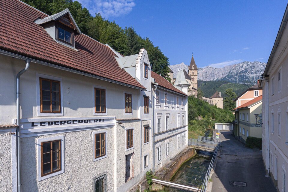 Ein malerisches Gebäude am Fluss, umgeben von grünen Hügeln. Im Hintergrund sind Berge und ein historischer Turm sichtbar. | © Verein Gerberei Salzer Museum