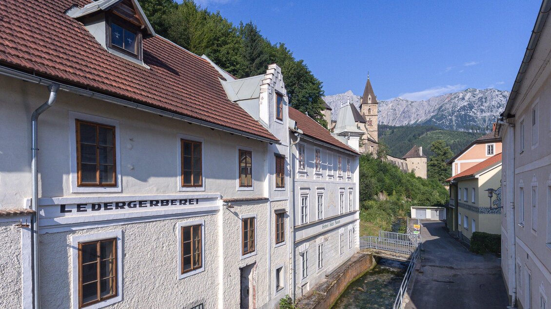 Ein malerisches Gebäude am Fluss, umgeben von grünen Hügeln. Im Hintergrund sind Berge und ein historischer Turm sichtbar. | © Verein Gerberei Salzer Museum