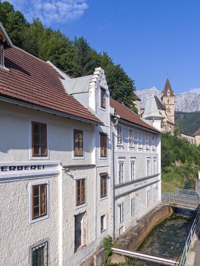 Ein malerisches Gebäude am Fluss, umgeben von grünen Hügeln. Im Hintergrund sind Berge und ein historischer Turm sichtbar. | © Verein Gerberei Salzer Museum