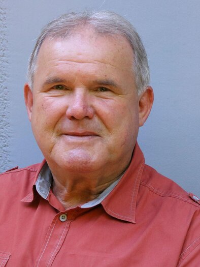 An older man with a friendly smile is sitting in front of a neutral background. He is wearing a red shirt and looks relaxed. | © Veronika Erhart