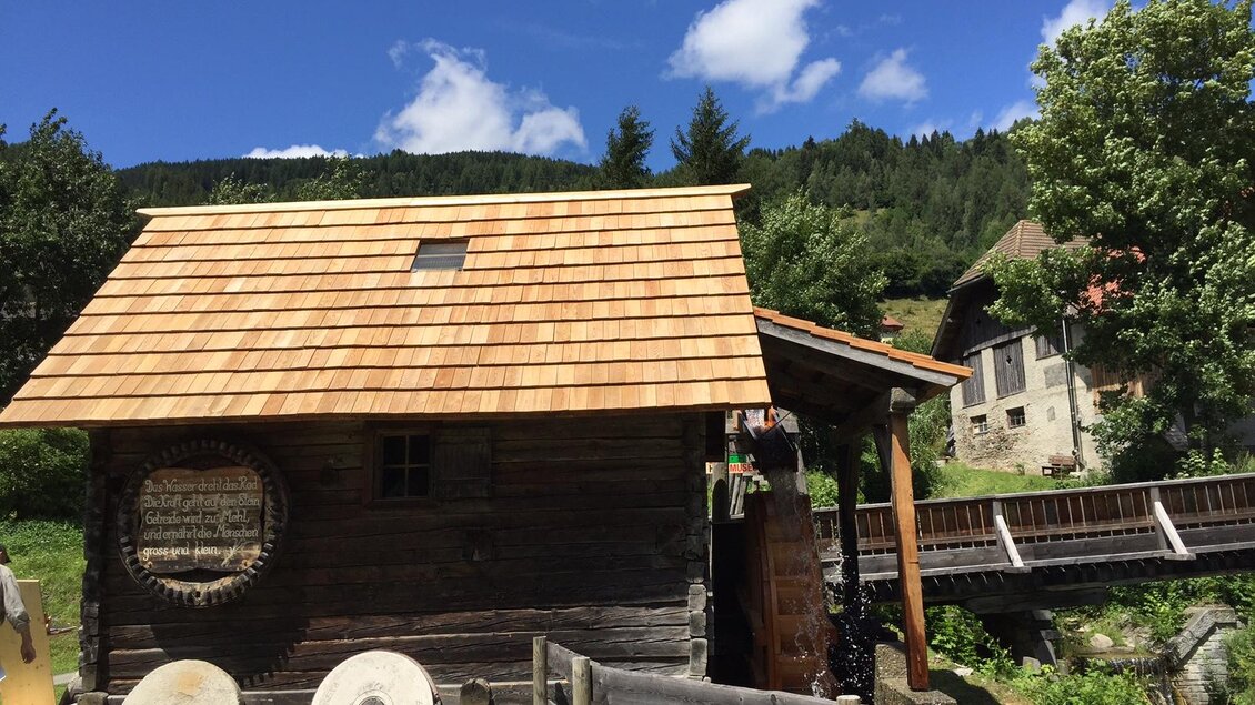 Ein rustikales Holzhaus mit einem Schindeldach steht in einer grünen Landschaft. Im Hintergrund sind Bäume und weitere Gebäude sichtbar, unter einem klaren blauen Himmel.