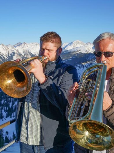 Zwei Männer spielen eine Trompete und ein Euphonium in den Bergen. Im Hintergrund sind schneebedeckte Gipfel und ein klarer Himmel zu sehen. | © Riesneralm Bergbahnen