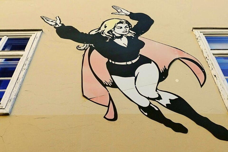 Eine Wandmalerei einer Superheldin, die durch die Luft fliegt. Sie trägt ein schwarzes Outfit und hat lange blonde Haare. | © Ruth Nezmahen