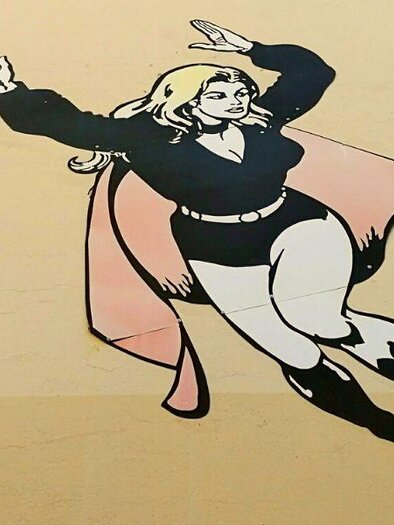 A mural of a superheroine flying through the air. She is wearing a black outfit and has long blonde hair. | © Ruth Nezmahen