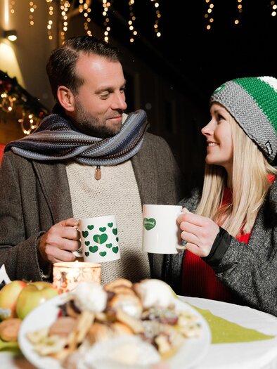 A couple stands at a festively decorated stall holding mugs in their hands. The atmosphere is cozy and Christmas-like, with lights in the background. | © Lupi Spuma