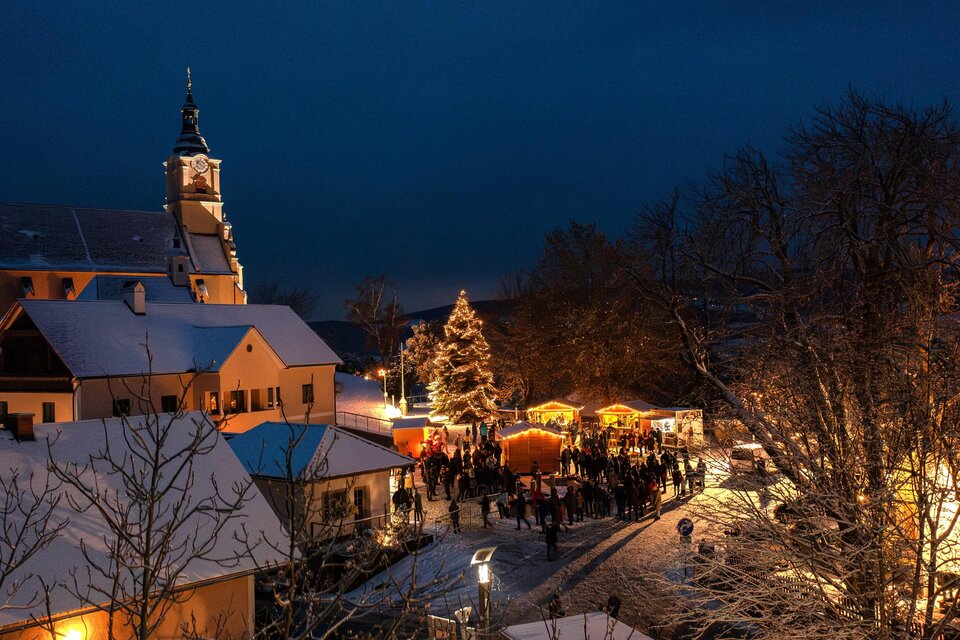 Eine winterliche Szene mit einem festlich beleuchteten Weihnachtsmarkt. Schnee bedeckt die Dächer, und Menschen versammeln sich um die Stände. | © Gemeinde Pöllauberg