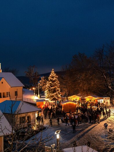 Eine winterliche Szene mit einem festlich beleuchteten Weihnachtsmarkt. Schnee bedeckt die Dächer, und Menschen versammeln sich um die Stände. | © Gemeinde Pöllauberg