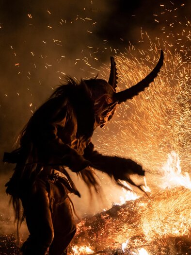 A dark scene with a spotted creature in a costume with horns. Sparks are flying from a fire in the background. | © Adobe Stock