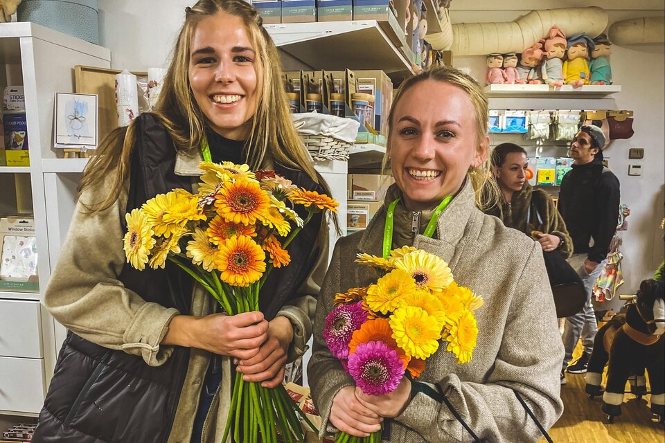 Zwei Frauen stehen in einem Geschäft und halten bunte Blumensträuße. Im Hintergrund sind andere Personen und Regale mit Waren zu sehen. | © Tourismusverband Oststeiermark
