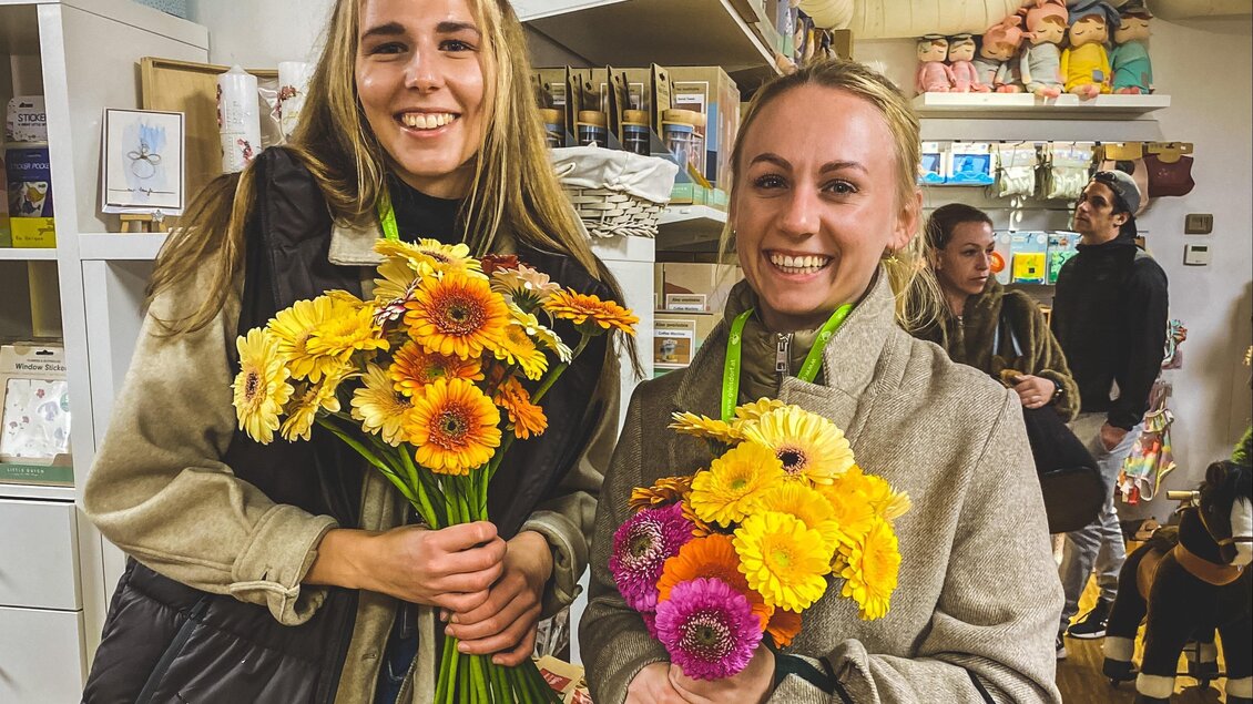Zwei Frauen stehen in einem Geschäft und halten bunte Blumensträuße. Im Hintergrund sind andere Personen und Regale mit Waren zu sehen. | © Tourismusverband Oststeiermark