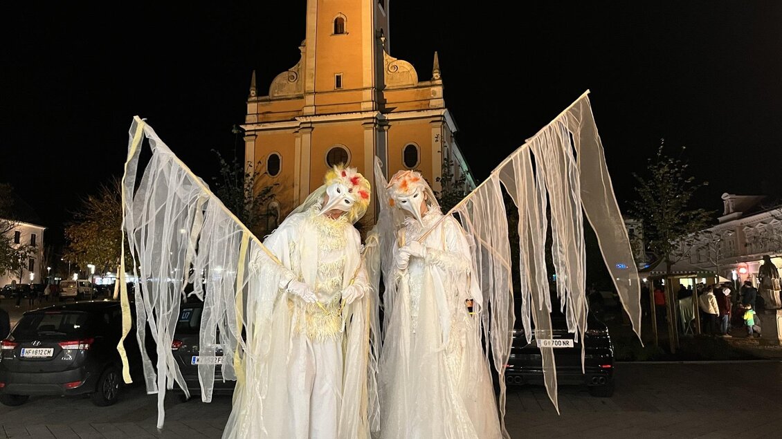 Zwei verkleidete Personen in weißen Kostümen stehen nachts vor einer gelben Kirche. Sie tragen Flügel aus Durchsichtigem Stoff und haben gruselige Masken auf. | © Tourismusverband Oststeiermark