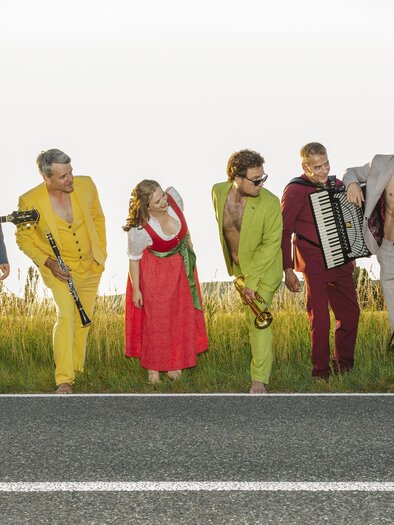 Als bunt gemischtes Sextett stehen die Global Kryner an der Landstraße - als würden sie Auto stoppen - die Sängerin im roten Dirndl und die Musiker in bunten Anzügen - von gelb über blau, grün, rot und grau - ohne Hemden und Socken und ohne Schuhe, in der Hand ihre Instrumente - Gitarre, Klarinette, Trompete, Akkordeon und Posaune. | © Oststeiermark Tourismus