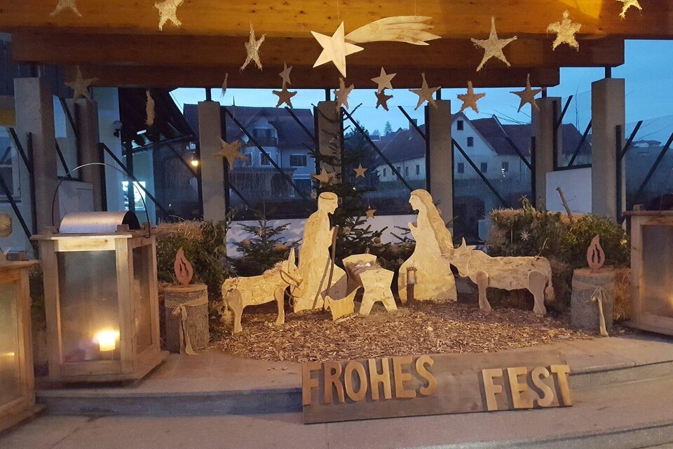 Eine stimmungsvolle Weihnachtskrippe mit Figuren von Maria, Josef und Tieren. Daneben steht der Schriftzug "FROHES FEST" und es leuchten Laternen im Hintergrund. | © Thermen- und Vulkanland Steiermark