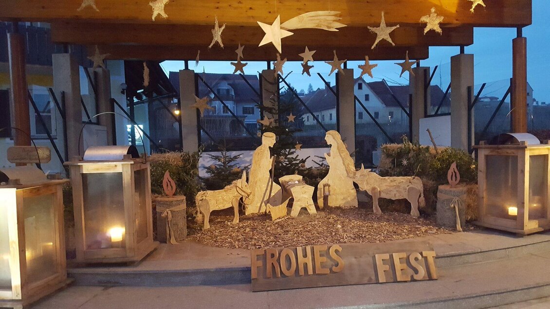Eine stimmungsvolle Weihnachtskrippe mit Figuren von Maria, Josef und Tieren. Daneben steht der Schriftzug "FROHES FEST" und es leuchten Laternen im Hintergrund. | © Thermen- und Vulkanland Steiermark