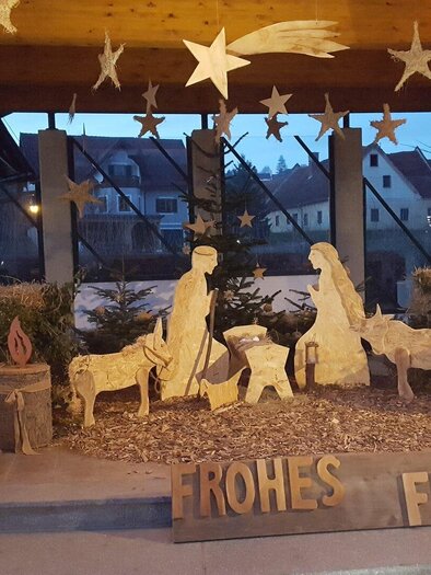 Eine stimmungsvolle Weihnachtskrippe mit Figuren von Maria, Josef und Tieren. Daneben steht der Schriftzug "FROHES FEST" und es leuchten Laternen im Hintergrund. | © Thermen- und Vulkanland Steiermark