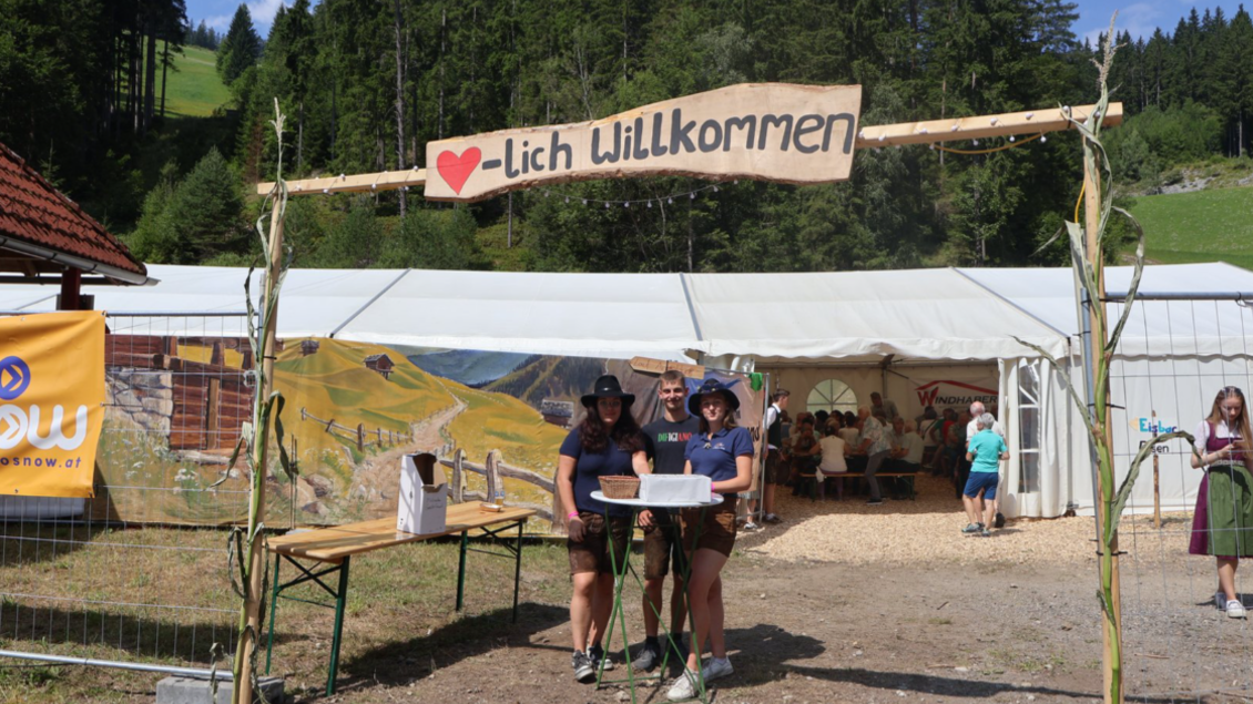 Eine Gruppe von Menschen steht unter einem Willkommensschild auf einem Festgelände in den Bergen. Im Hintergrund sind Zelte und eine malerische Landschaft mit Wäldern zu sehen. | © LJ Langenwang