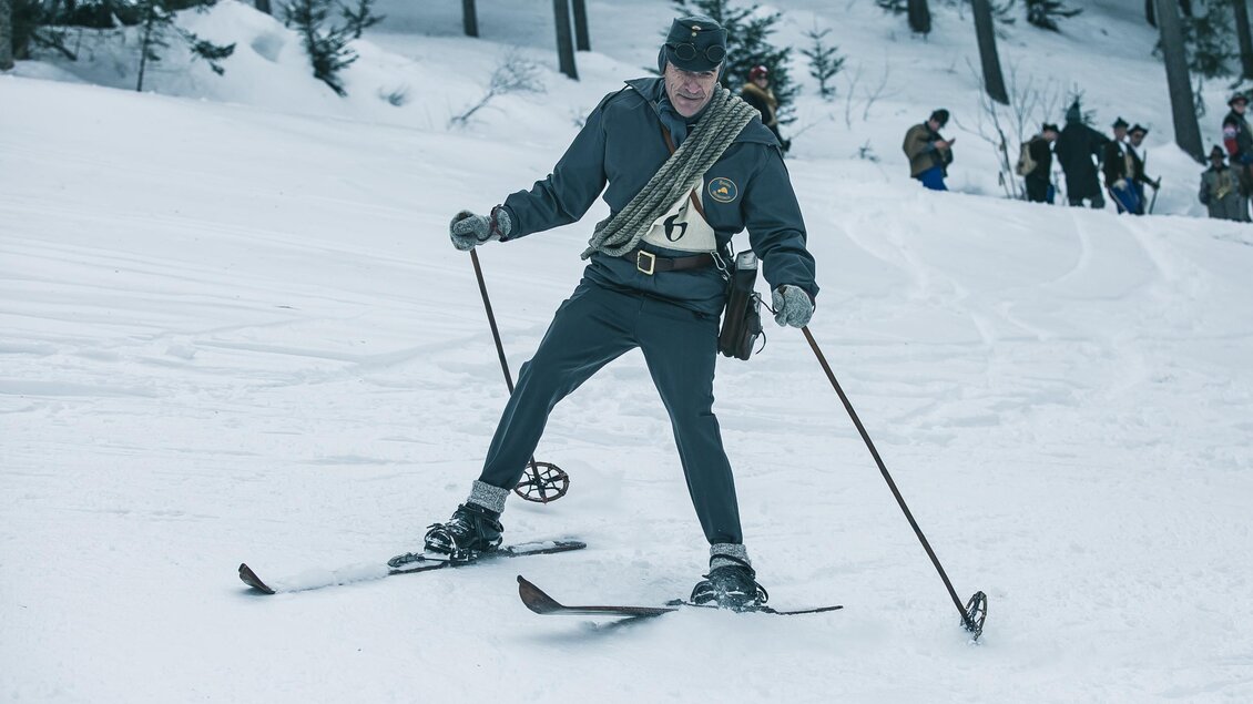 Ein Skifahrer in einer historischen Uniform fährt durch den Schnee. Im Hintergrund sind Menschen sichtbar, die zuschauen. | © Oliver Königshofer
