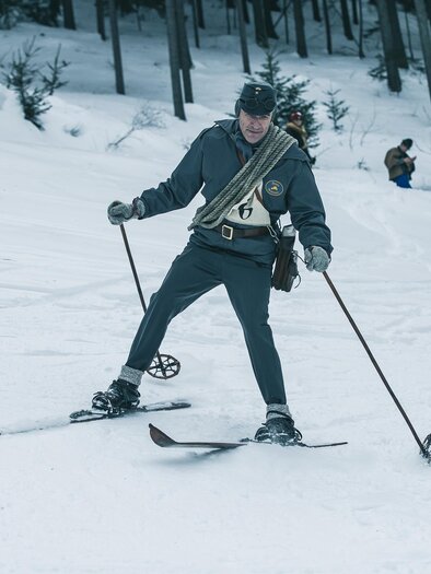 A skier in a historical uniform is skiing through the snow. In the background, people are visible watching. | © Oliver Königshofer