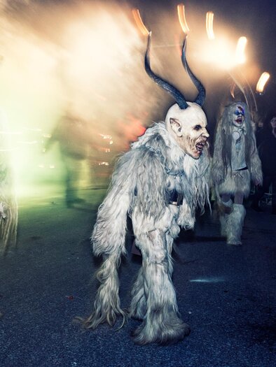 A group of warrior-like figures in furry costumes with horns moves through a festive atmosphere. In the background, people and light displays can be seen. | © M. Hierzer